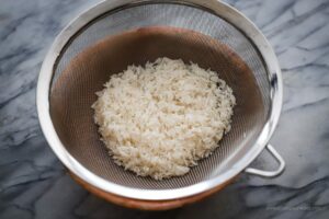 Image of uncooked rinsed jasmine rice in mesh strainer.