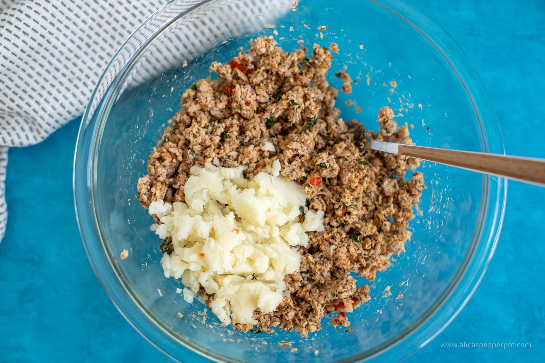 Mashed potato with seasoned salmon cake mixture in a clear mixing bowl.