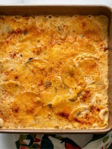 A golden-brown scalloped eddoe dish baked in a square pan, topped with spices, resting on a colorful floral kitchen towel.