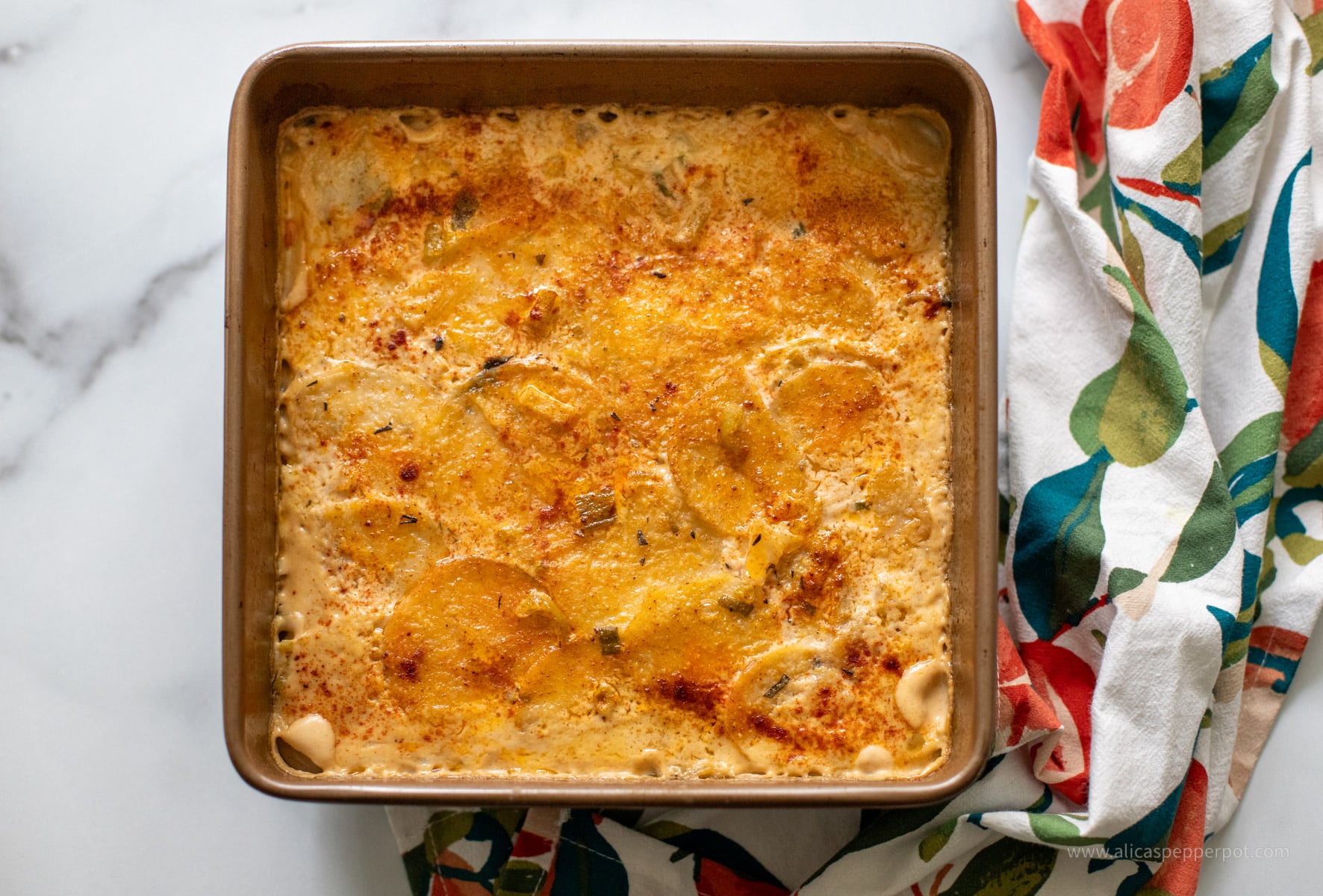 The image shows a delicious scalloped eddoe dish baked in a square metal pan. The golden brown top is beautifully crisp with a light dusting of paprika or cayenne pepper, adding a vibrant color. You can see thinly sliced eddoes. A colorful kitchen towel with floral patterns is positioned next to the pan.