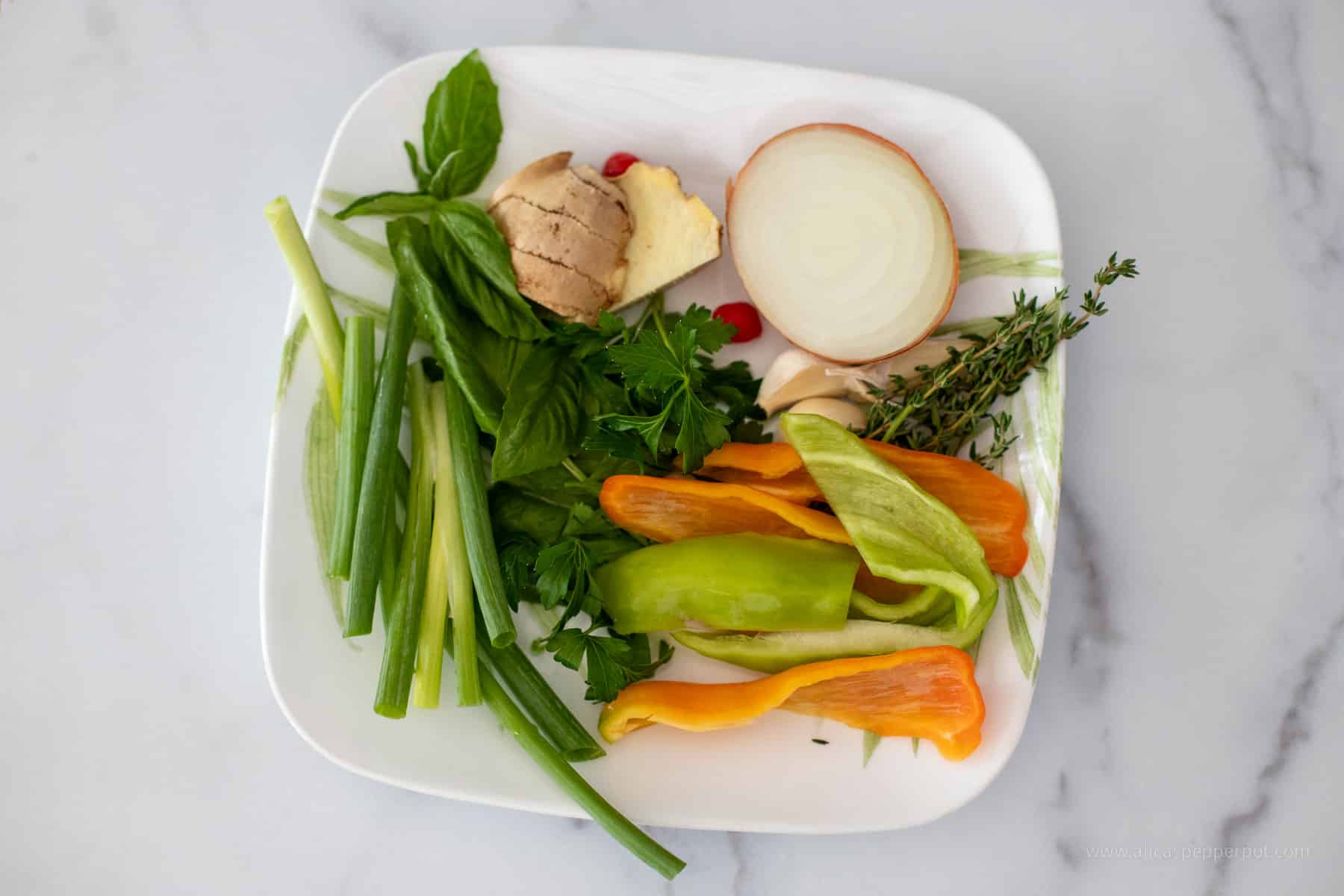 A white square plate with aromatics such as scallion ginger, pepper, onion, garlic, cubanelle peppers, basil, and thyme