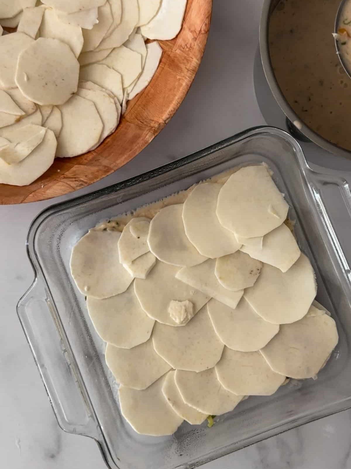 Thinly sliced eddoes layered with coconut milk sauce. One bowl has sliced eddoes, a pot with the coconut milk is on the side.