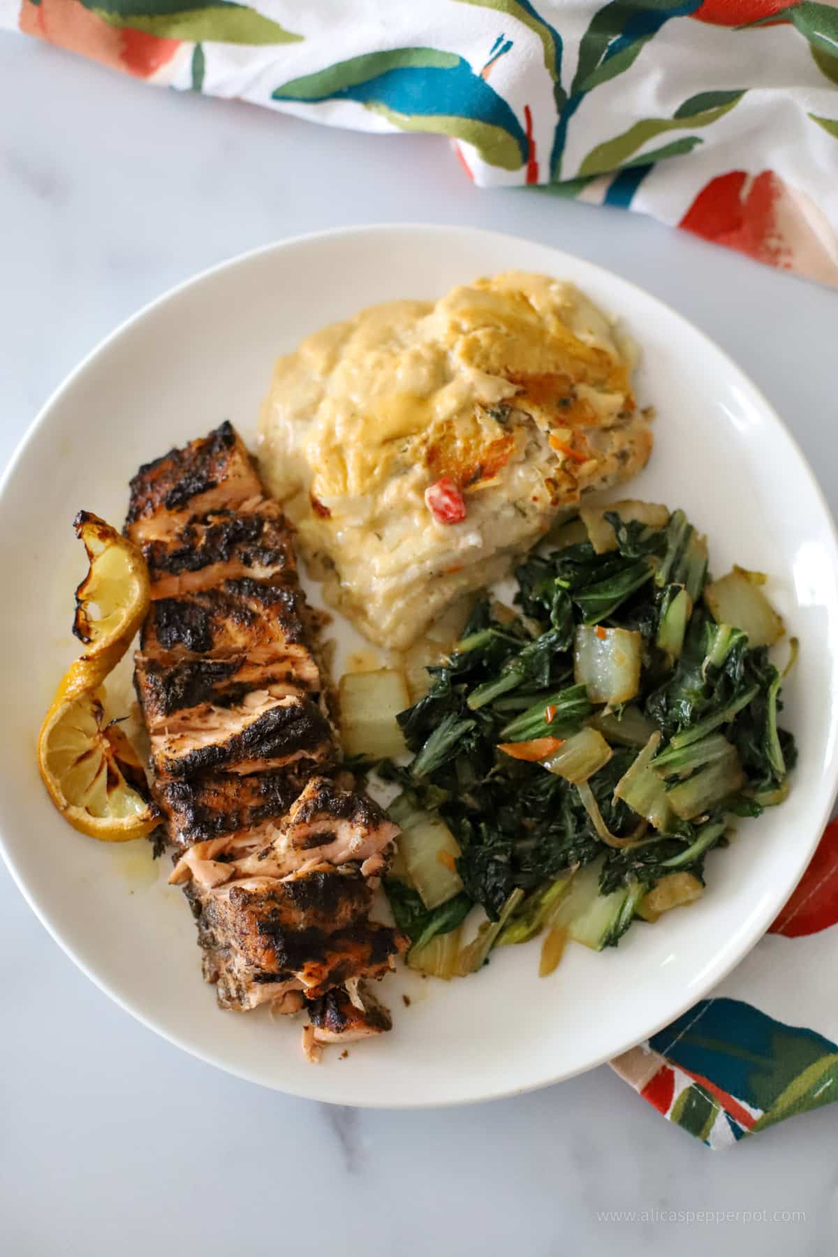 A dinner plate with Jerked salmon, scalloped eddoes, and sauteed bok choy served in a white plate with a floral napkin on the side.