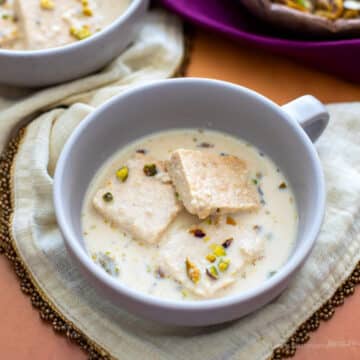 Rasmalai made with ricotta cheese in a grey dessert bowl on an ivory napkin. There are pistachios and almonds for toppings in a wooden bowl in the background.